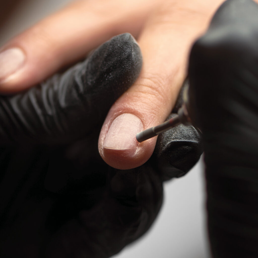 Technicienne ongulaire appliquant un produit au pinceau sur l’ongle naturel d’une cliente en salon.