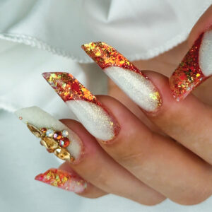 Pose ongulaire stiletto avec encapsulation de paillettes orangées et décoration strass en relief sur base blanche nacrée.